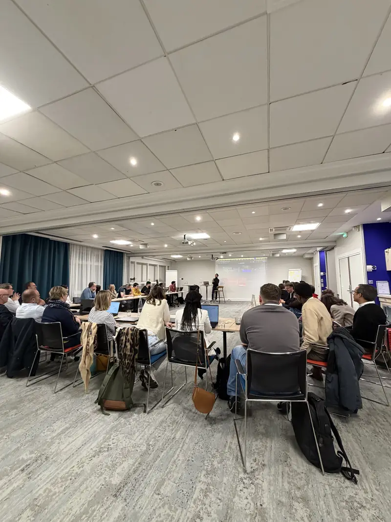 Une grande salle de formation vue du fond : des dizaines de participants de dos, face à l'écran et à l'intervenant.