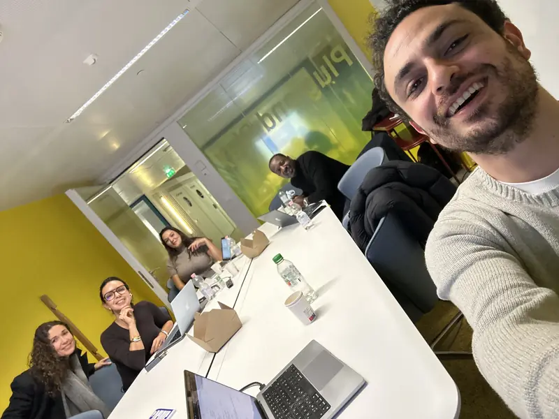 Paul Slama en selfie avec quatre participants assis autour d'une table, ordinateurs ouverts, dans une salle aux murs jaunes.